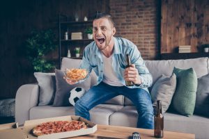 man celebrating on the couch at home while enjoying chips. beer, and a pizza