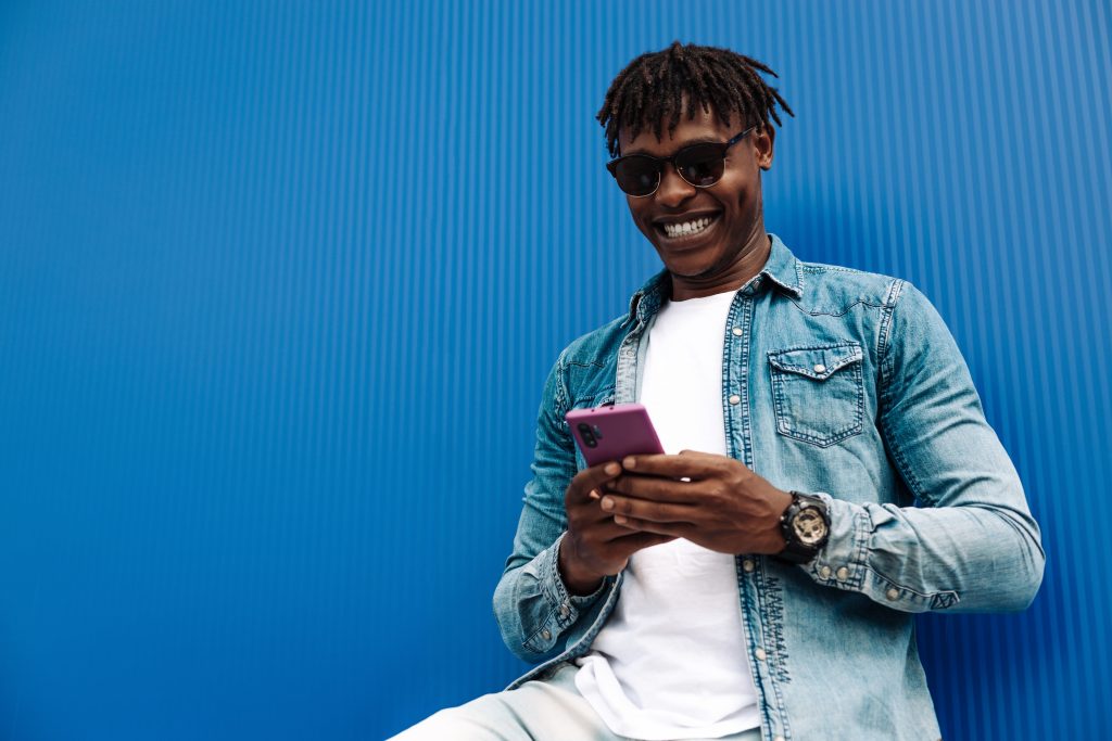 Young black guy with sunglasses smiling looking at phone against bright blue background