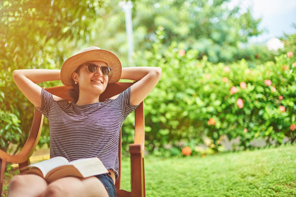 Young woman in sunglasses and straw hat leaning back in lawn chair with book on lap