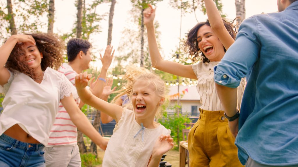 Family and friends dancing outside with laughing little blonde girl in front