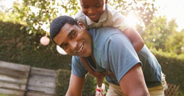 Young Black Boy Playing On Dad Back In A Garden