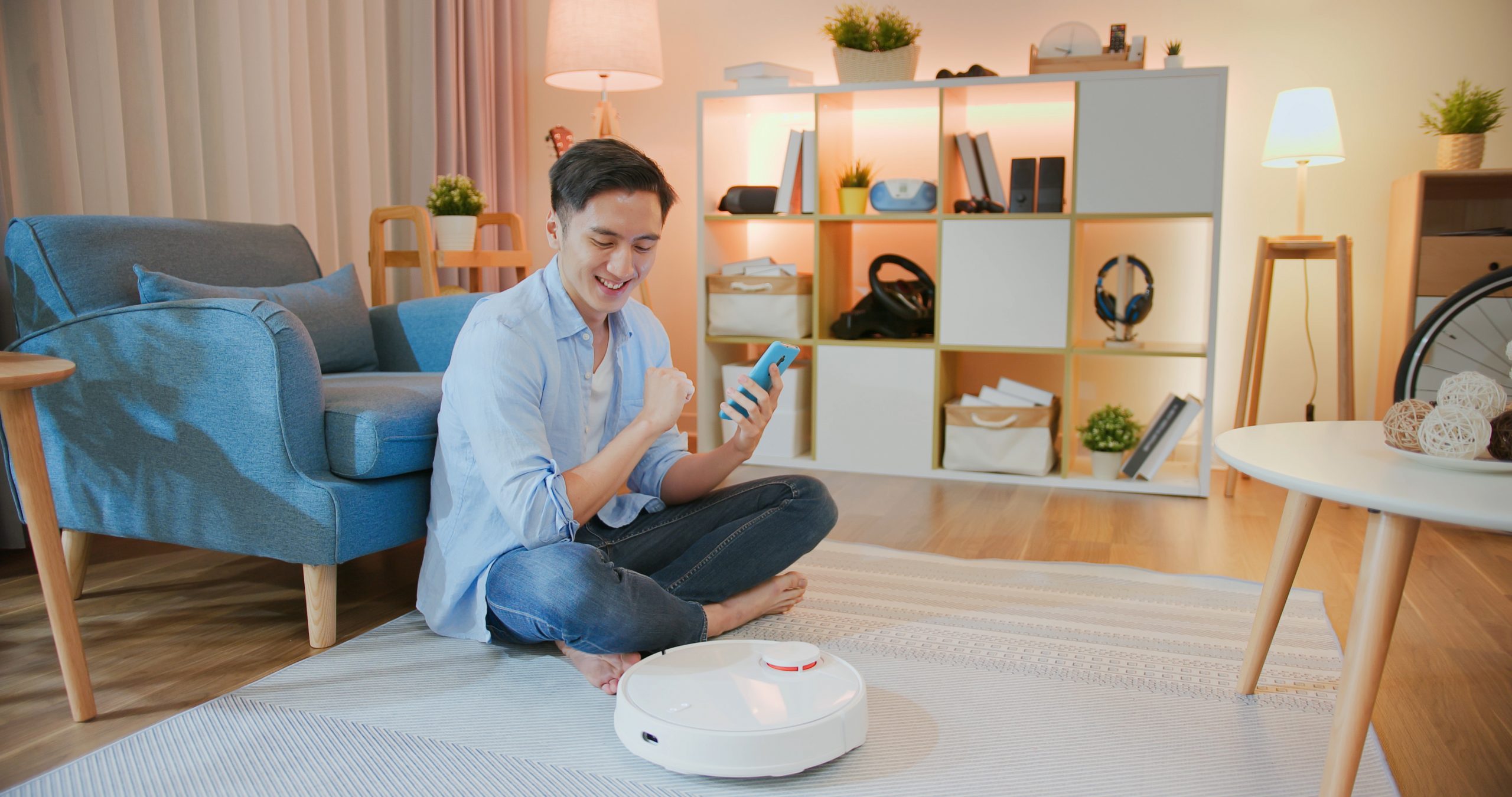Young Asian man sitting on floor programming robot vacuum with smartphone