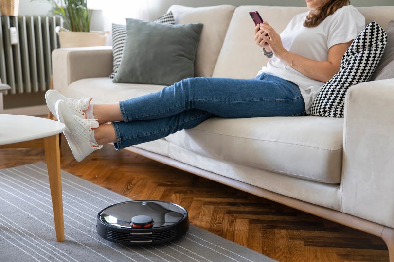 Young caucasian woman sitting on sofa with feet up, robot vacuum working nearby