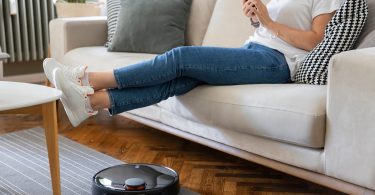 Young caucasian woman sitting on sofa with feet up, robot vacuum working nearby