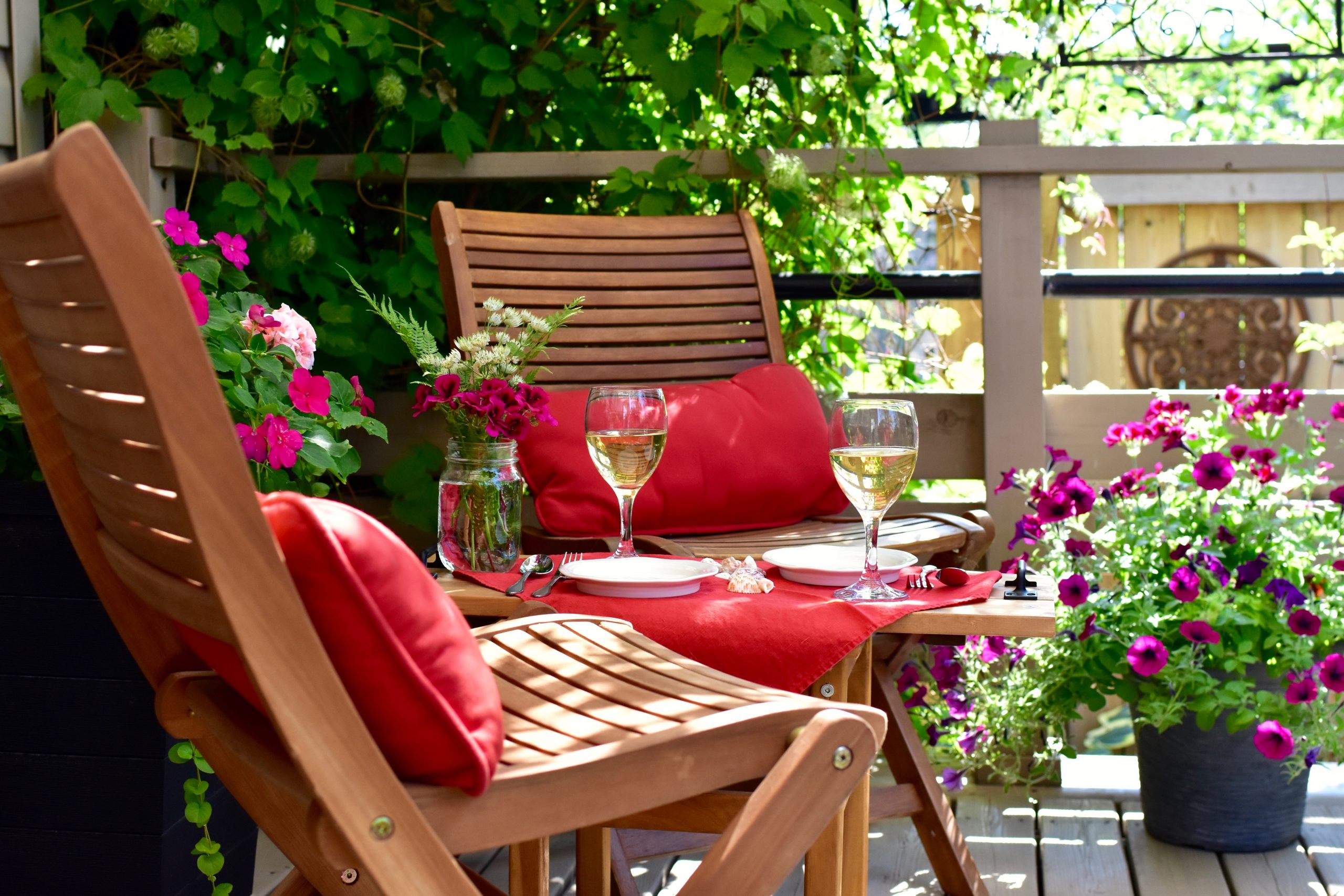 Wood patio chairs with red cushions, pretty flowers nearby, wine on the table