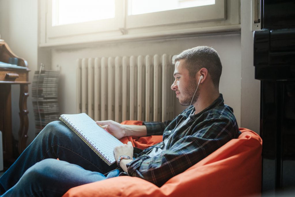 College-age white man relaxing on beanbag with earphones reading notebook