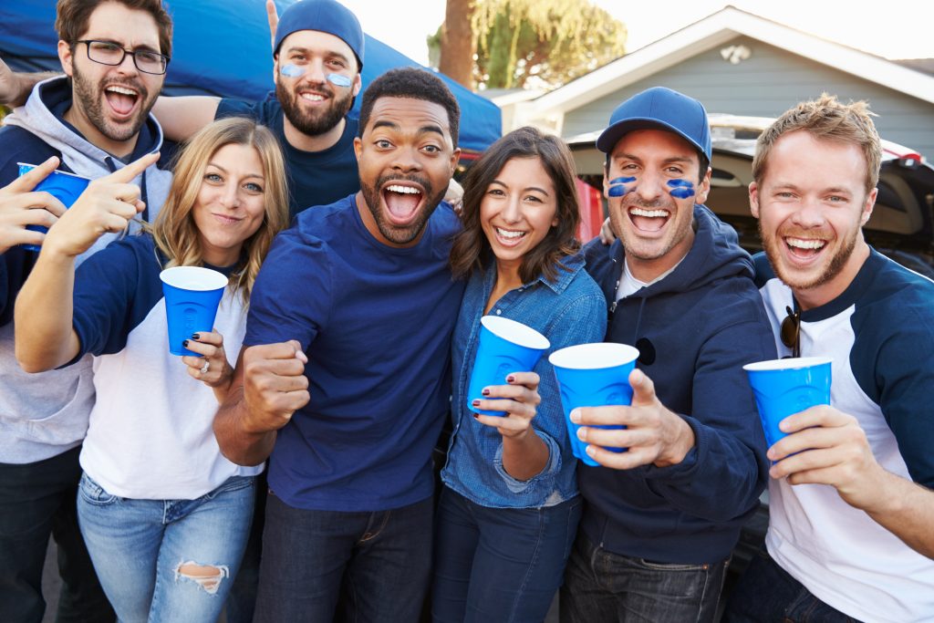 Seven young adult tailgaters in blue team gear big smiles holding blue cups