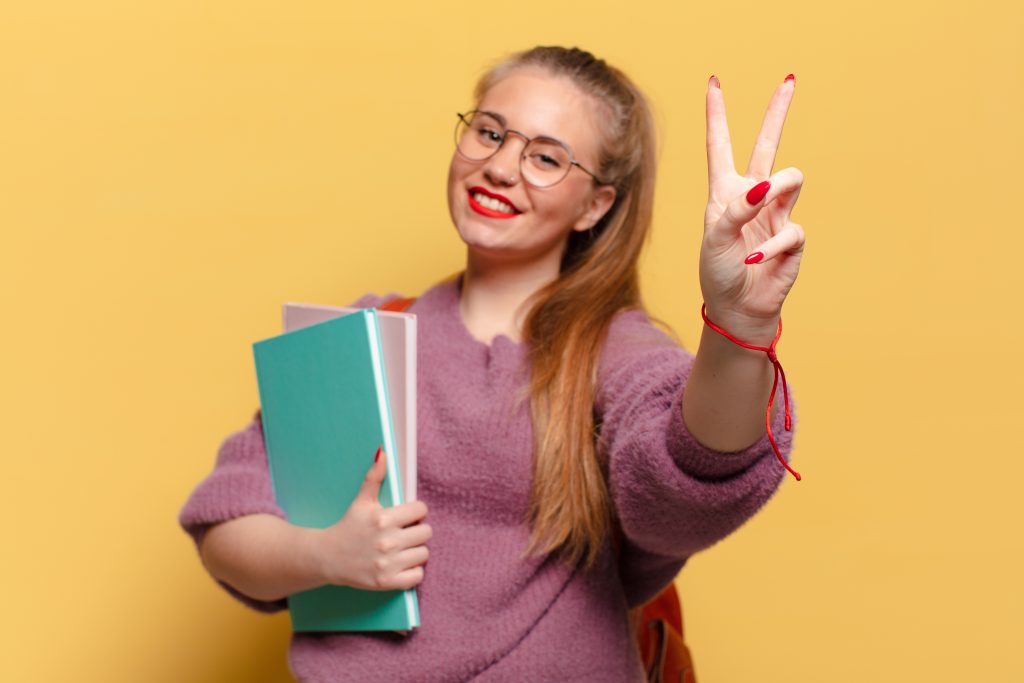 White high-school girl with long blonde ponytail and glasses holding books and giving peace sign