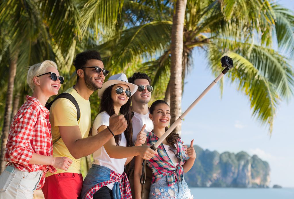 Group of diverse young adults in island location, all in sunglasses, taking picture with selfie stick