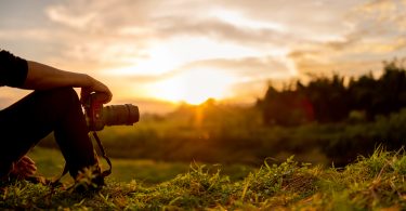 unset photo with just inset shot of young man's arm propped on bent knee holding camera with long lens, sitting, looking at sunset