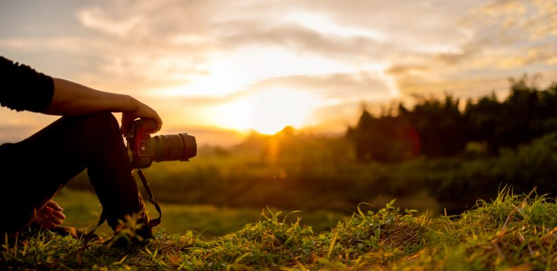 unset photo with just inset shot of young man's arm propped on bent knee holding camera with long lens, sitting, looking at sunset