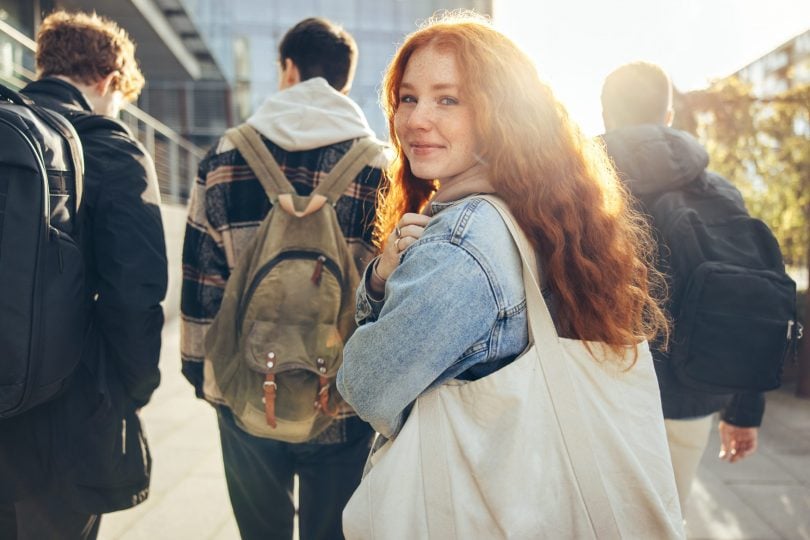 Female student with long wavy red hair with shoulder bookbag glancing back