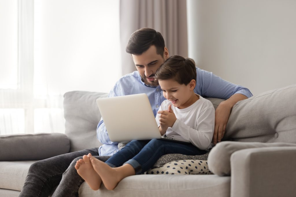 Hispanic young father and son sitting on sofa looking at laptop together